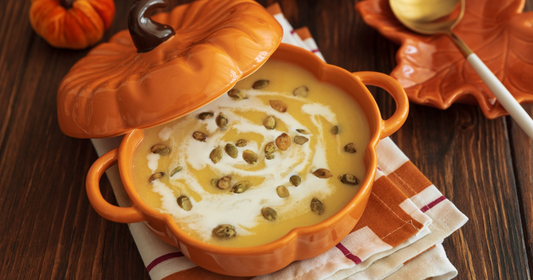 Soup in a pumpkin shaped pot with silverware on the side. 
